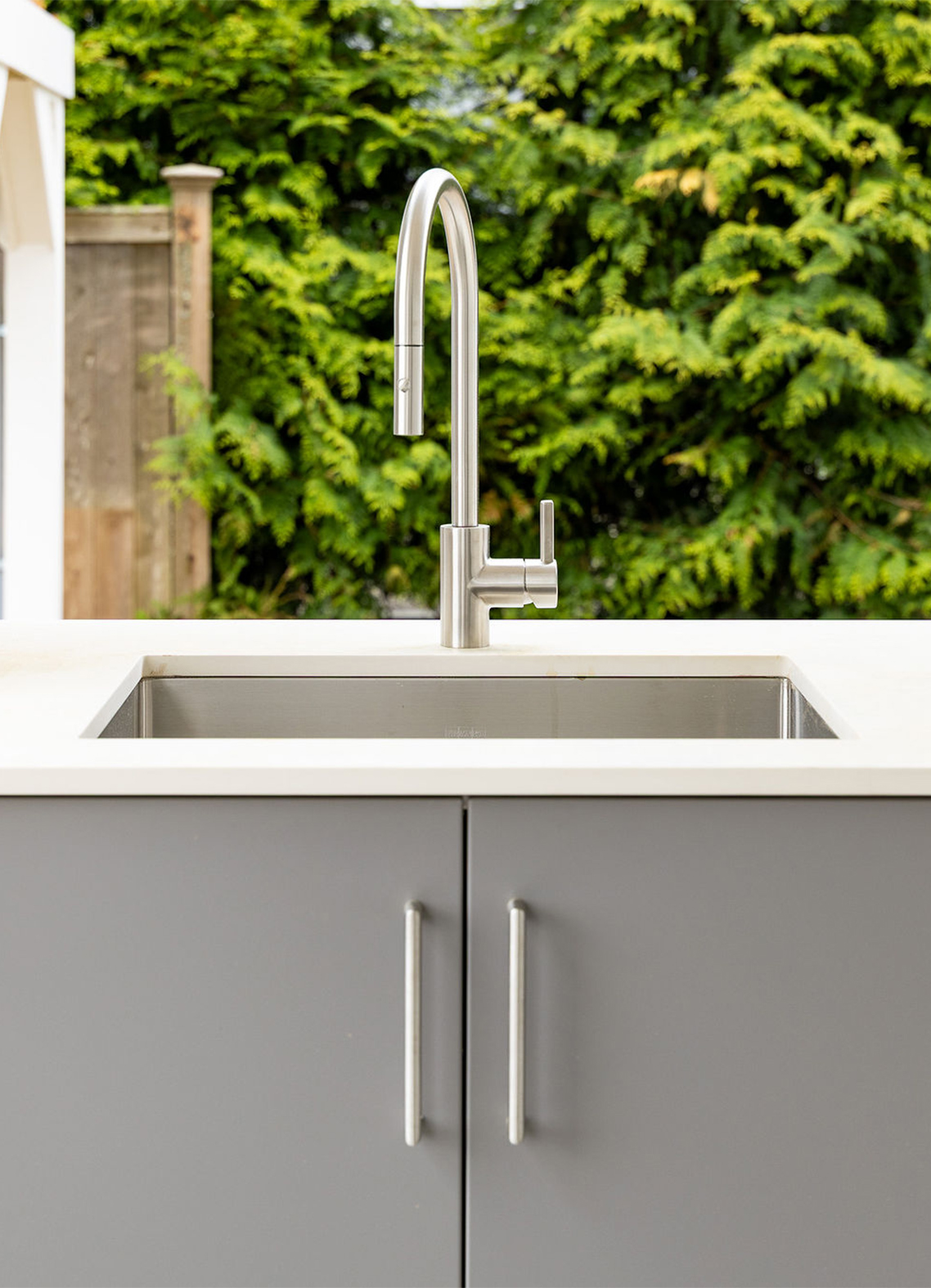 Outdoor stainless steel kitchen faucet over a sink on a gray cabinet base, with a green hedge in the background.