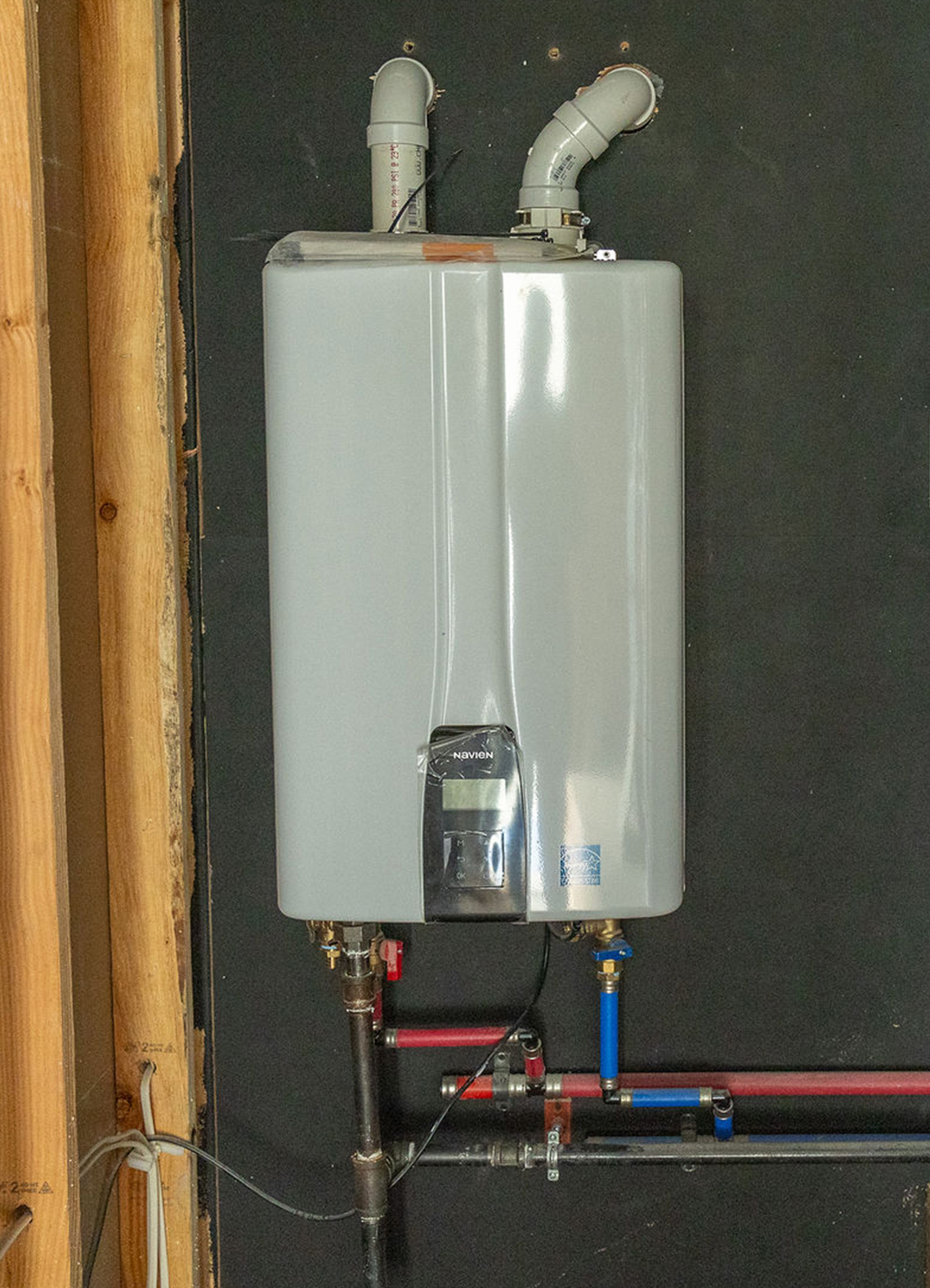 Wall-mounted Navien tankless water heater on a dark wall, with white PVC vent pipes above and red/blue water supply lines below.